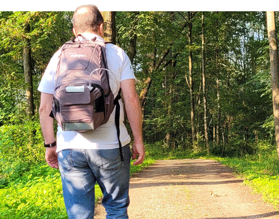Person von hinten mit einem tragbaren Sauerstoffgerät im Rucksack, geht durch eine Naturlandschaft, symbolisiert mobile Freiheit.